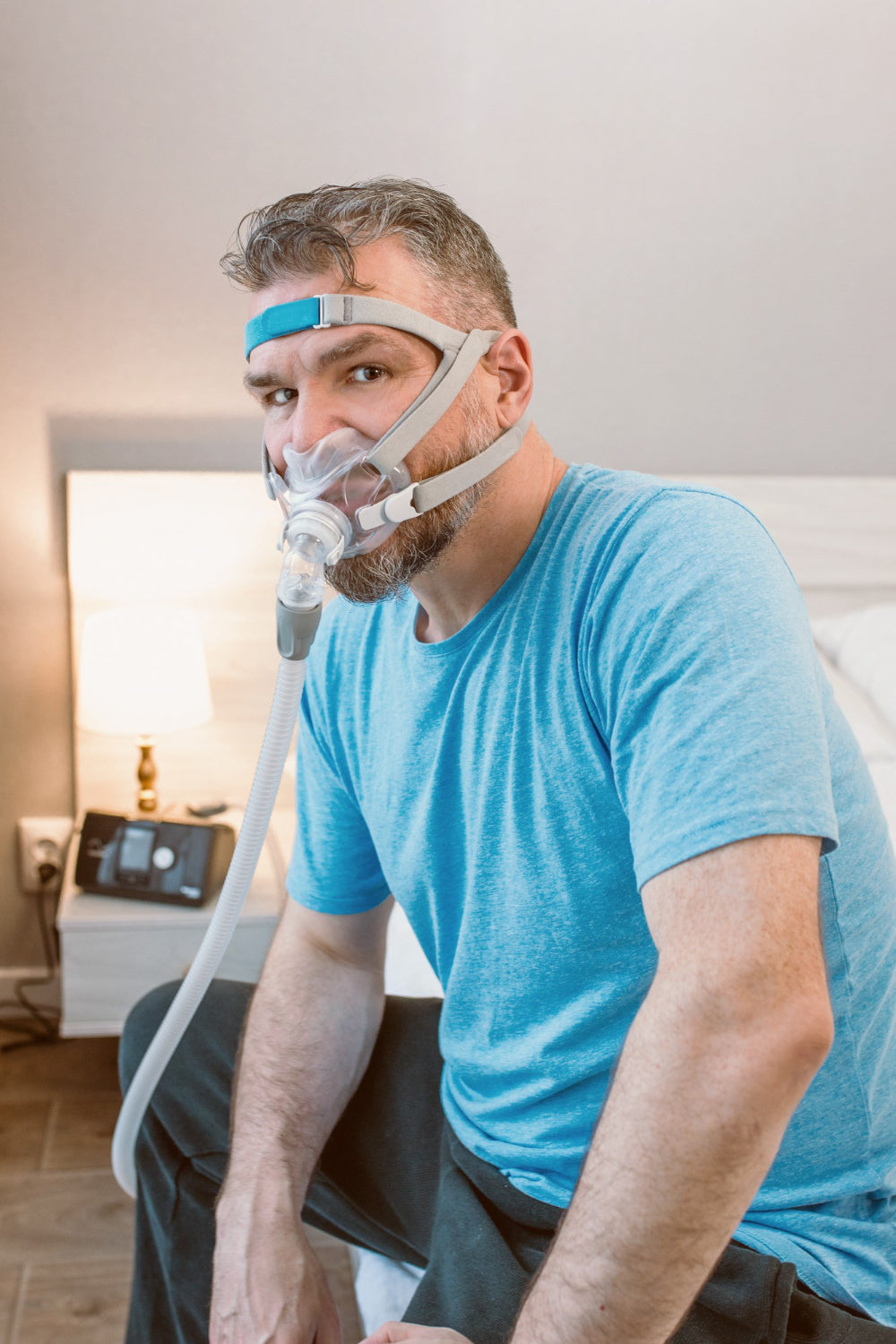 Image: A man sitting on the edge of a bed wearing a CPAP mask and a scowl on his face.