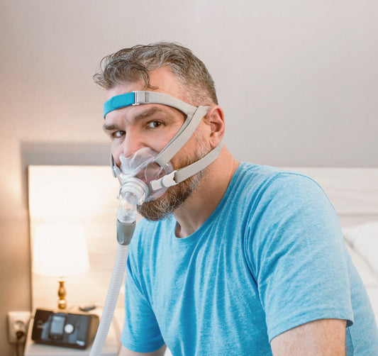 Image: A man sitting on the edge of a bed wearing a CPAP mask and a scowl on his face.