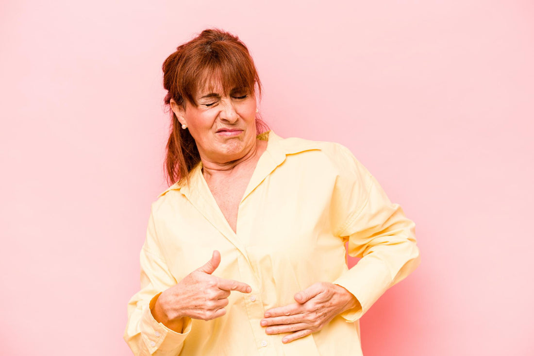 Image: Woman wearing a yellow shirt and pointing to her belly while making a face that looks like she is in pain.