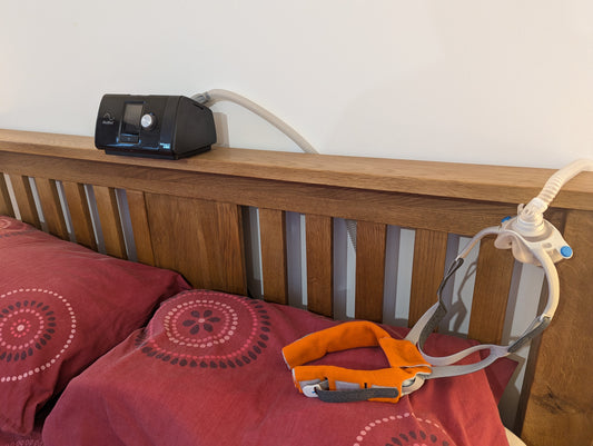 Image: A CPAP machine sitting on a bed headboard with a Resmed F40 CPAP mask and Stag's Head CPAP Strap Cover laying on the pillow.