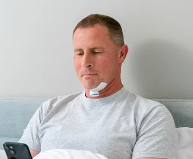 Image: A man sitting in bed looking at his phone while wearing a Nyxoah Genio sleep apnea device