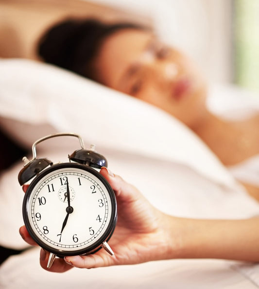 Image: A woman lays her head a pillow in a blurred background, with an old-fashioned alarm clock in the foreground.